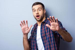 Terrified with something. Frustrated young man in casual shirt keeping mouth open and looking terrified while standing against grey background photo