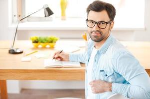 Focused on success. Handsome young man in shirt and eyewear writing in note pad and looking at camera over shoulders while sitting at his working place photo