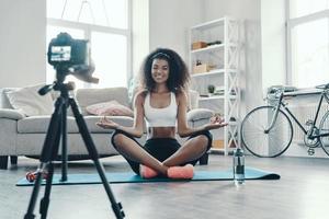 Attractive young African woman sitting in yoga position and smiling while making social media video photo