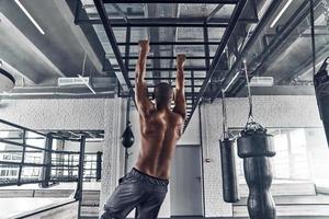 On the way to great shape. Rear view of shirtless young African man doing pull-ups while exercising in the gym photo