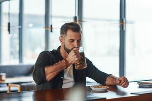 Handsome young man in casual clothing drinking beer while spending time in the pub photo