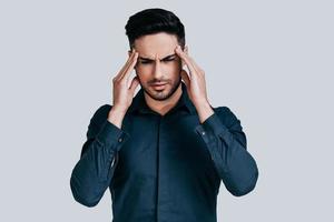 Terrible headache. Frustrated young man touching his head with hands and making face while standing against grey background photo