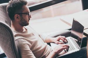 Working with pleasure. Side view of handsome man using his laptop while sitting in chair in front of window photo