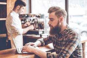 Working in cafe. Side view of young handsome bearded man using his laptop while sitting at bar counter at cafe with barista at the background photo