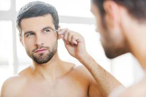 Tweezing his eyebrows. Handsome young man tweezing his eyebrows and looking at himself while standing in front of the mirror photo