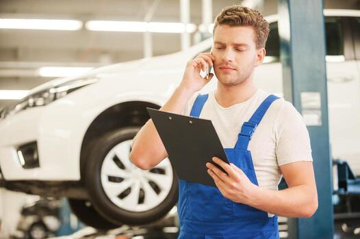 Discussing some car problems. Handsome young man talking on the mobile phone and looking at clipboard while standing in workshop with car in the background photo