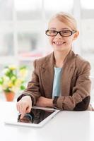 Making business easier. Cheerful little girl in glasses and formalwear sitting at the table and working on digital tablet photo