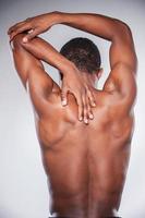 Joint pain. Rear view of young shirtless African man touching his neck and elbow while standing against grey background