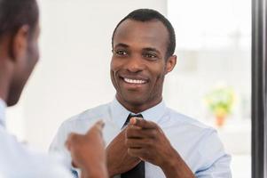 Feeling confident. Young African man adjusting his necktie and pointing himself at the mirror photo