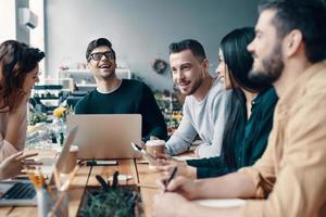 Happy to work together. Group of young modern people in smart casual wear discussing something and smiling while working in the creative office photo