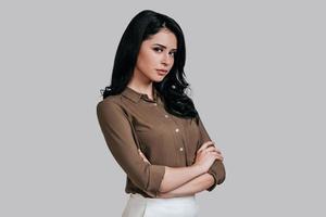 Modern woman. Beautiful young woman in smart casual wear keeping arms crossed and looking at camera while standing against grey background photo