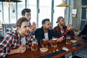 Cheering young men in casual clothing drinking beer and watching sport game while sitting in the pub photo