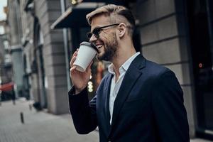 Simply happy. Handsome young man in smart casual wear drinking coffee and smiling while standing outdoors photo