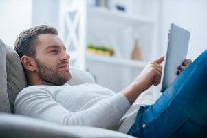 Enjoying his leisure time at home. Side view of handsome young man working on digital tablet and looking relaxed while lying on the couch at home photo