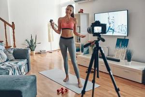 toda la longitud de una hermosa joven con ropa deportiva haciendo ejercicio y sonriendo mientras hace videos en las redes sociales foto