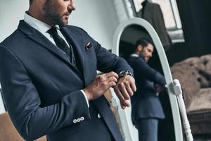Am I late Close up of young man in full suit checking the time while standing in front of the mirror indoors photo