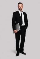 Pure confidence. Full length of handsome young man in full suit carrying laptop and looking at camera while standing against grey background photo