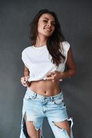 chica irresistible. atractiva mujer joven ajustando su camiseta y sonriendo mientras está de pie contra el fondo gris foto