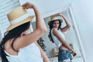 Trendy girl. Beautiful young woman looking at her reflection in the mirror while trying on her sun hat photo