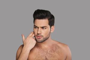 What is wrong with my skin Confused young man looking away and touching his face with hand while standing against grey background photo