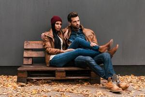 Enjoying time together. Beautiful young couple bonding to each other while sitting on the wooden pallet with grey wall in the background and fallen leaves on the floor photo