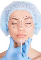 Lips improvement. Attractive young woman in medical headwear and sketches on face keeping eyes closed while doctors hand making an injection in her lips photo