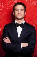 Handsome in bow tie. Handsome young man in suit and bow tie keeping arms crossed and looking at camera while standing against red background photo