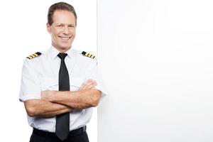 Confident and experienced pilot. Confident male pilot in uniform leaning at the copy space and smiling while standing against white background photo