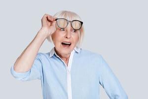 increíble. sorprendida anciana ajustando los anteojos y haciendo una mueca mientras se enfrenta a un fondo gris foto