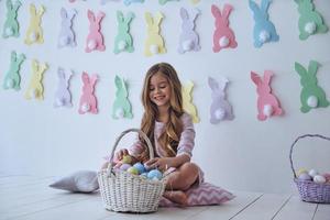 sintiendo alegría por las vacaciones. linda niñita jugando con canasta de pascua y sonriendo mientras se sienta en la almohada con decoración en el fondo foto