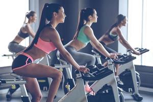 Beautiful ride. Side view of young beautiful women with perfect bodies in sportswear looking away with smile while cycling at gym photo