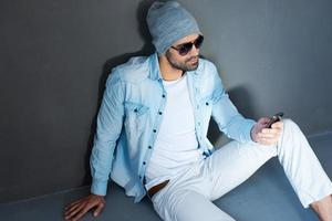 Texting to friend. Top view of handsome young man in eyewear holding mobile phone while sitting on the floor against grey background photo