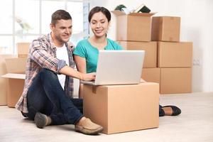 Surfing the net together. Cheerful young couple sitting on the floor and working on laptop while cardboard boxes laying on the background photo