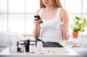 Consulting with doctor. Cropped image of sick woman holding a mobile phone and business card while sitting in bed at her apartment with medicines on the foreground photo