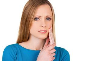 Suffering from toothache. Frustrated young woman touching her cheek and looking at camera while standing isolated on white photo