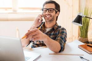 Creative worker. Cheerful young man talking on mobile phone and pointing at the laptop while sitting at his working place photo