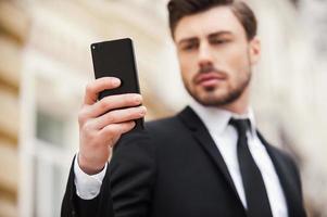 Always connected. Confident young man in formalwear holding mobile phone while standing outdoors photo