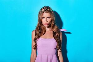 Boredom. Disappointed young woman in tiara looking at camera while standing against blue background photo