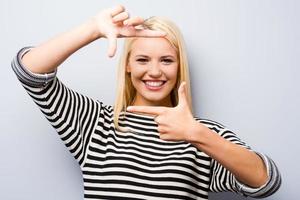 marco de dedo gesticular. hermosa joven rubia mirando a la cámara y haciendo gestos con el marco del dedo mientras se enfrenta a un fondo gris foto