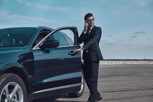 experto en negocios confiado. toda la longitud de un apuesto joven hombre de negocios hablando por teléfono mientras entra en su auto al aire libre foto