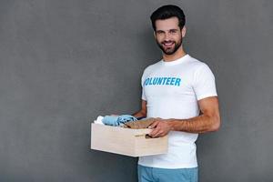 disfrutando retribuir a la comunidad. joven confiado con camiseta de voluntario sosteniendo una caja de donación en sus manos y mirando la cámara con una sonrisa mientras se enfrenta a un fondo gris foto