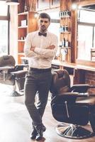 Barber expert. Full length of young handsome man looking at camera and keeping arms crossed while standing at barbershop photo