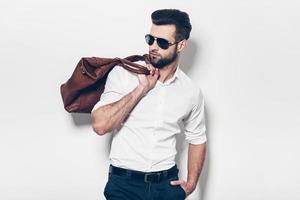 Ready to go. Handsome young man in white shirt carrying leather bag on shoulder and looking away while standing against white background photo