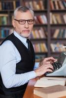 Confident author. Side view of confident grey hair senior man in formalwear sitting at the typewriter and looking at camera with bookshelf in the background photo