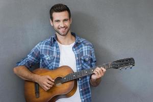 Young man with guitar. Happy young man playing the guitar and looking at camera while standing against grey background photo