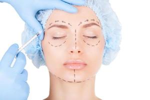 Botox injection. Attractive young woman in medical headwear and sketches on face keeping eyes closed while doctors hand making an injection in face photo