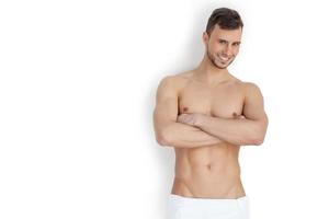 Keeping his body in good shape. Cheerful young muscular man looking at camera and keeping arms crossed while standing isolated on white background photo