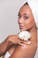 Natural beauty. Beautiful young Afro-American shirtless woman holding flower on her shoulder and looking at camera while isolated on gray background
