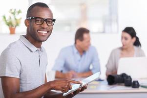 Cheerful team leader. Handsome young African man working on digital tablet and smiling while two people working on background photo