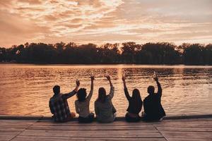 disfrutando de la hermosa puesta de sol. vista trasera de jóvenes con ropa informal brindando con botellas de cerveza mientras se sientan en el muelle foto